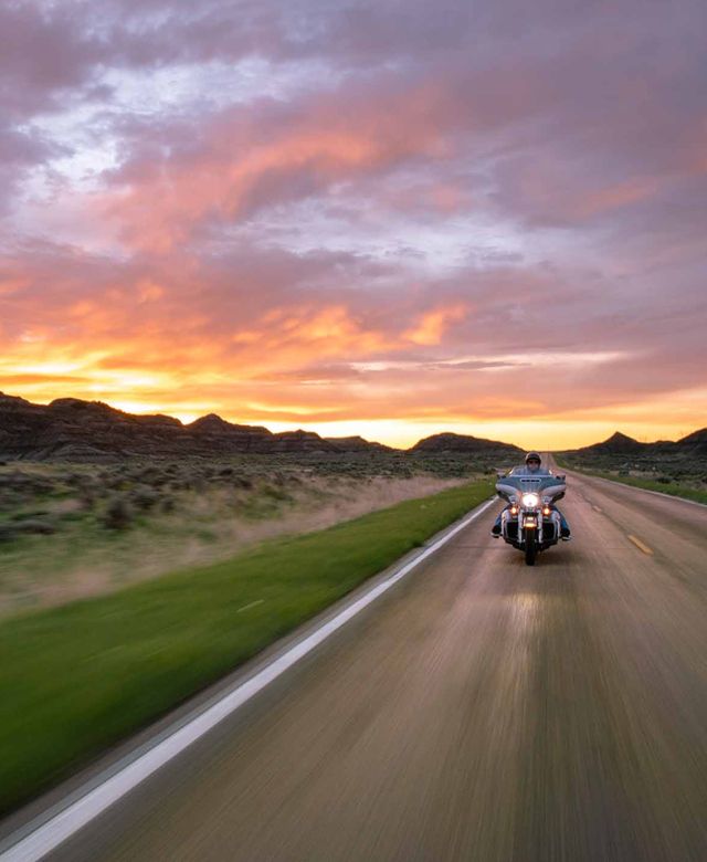 Motorcyclist crusing down a two lane highway in the Terry Badlands, with a purple and gold sunset in the background