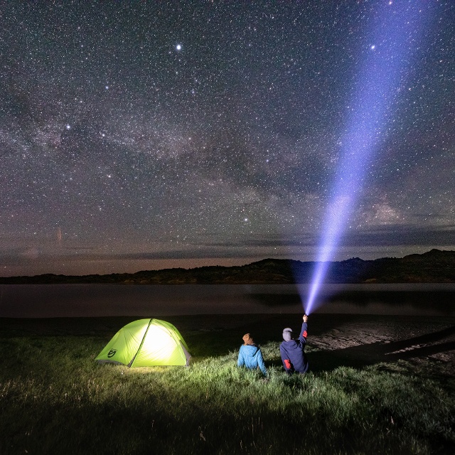 A star filled night sky. Two people sit in the foreground next to their tent.